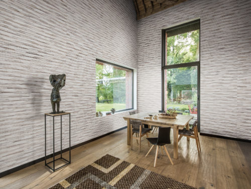 Pièce haute de plafond avec d'immenses fenêtres laissant entrer l'extérieur. Plus de nature sur les murs avec le revêtement mural Omexco Shades of Pale avec de l'abaca et du cuir simili.