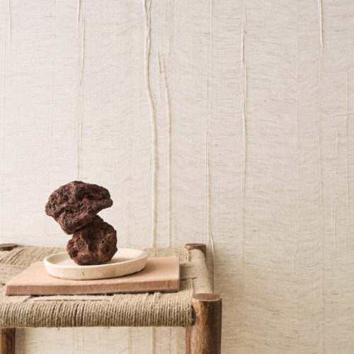 Une scène soigneusement composée mettant en avant la collection Pure d’Omexco, référence PU341, avec du lin tissé et plié avec une sensibilité artistique. La texture plissée du revêtement mural introduit un subtil jeu de lumière et d’ombre, créant un arrière-plan raffiné mais tactile. La composition comprend un tabouret rustique en bois avec un siège en corde tissée, surmonté d’un plat en céramique contenant deux roches volcaniques empilées, combinant des éléments naturels bruts à un artisanat raffiné. Les tons neutres et les matériaux naturels créent une atmosphère sereine et organique qui s’harmonise parfaitement avec les intérieurs minimalistes et artisanaux.