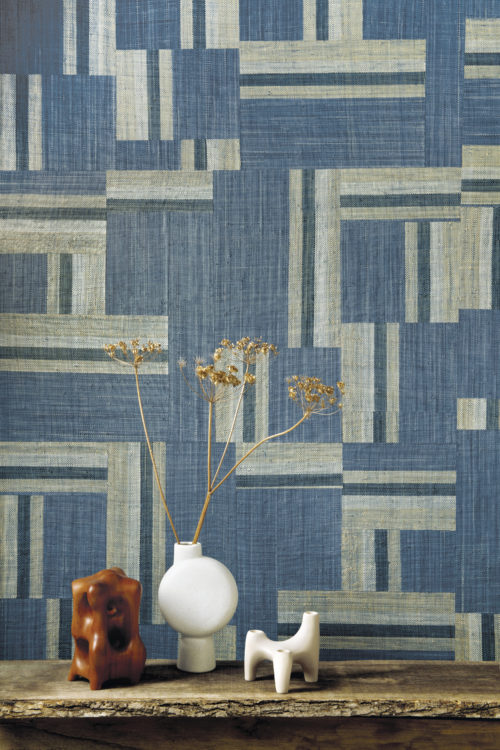 L'artisanat authentique incarne l'audacieuse collection Atelier conçue par les esprits créatifs d'Omexco. Cette image montre un impressionnant patchwork bleu denim en raphia tissé à la main. Devant le mur, nous voyons une table d'appoint en bois avec des éléments décoratifs en céramique blanche et brune et des fleurs séchées.