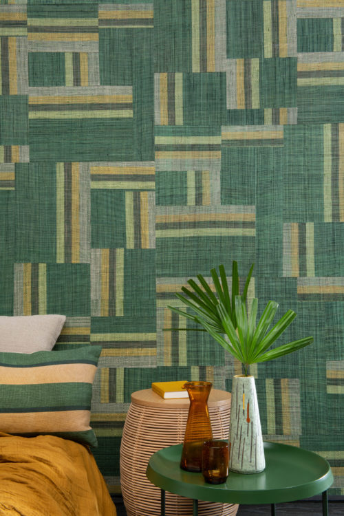 L'artisanat authentique incarne l'audacieuse collection Atelier conçue par les esprits créatifs d'Omexco. Cette image montre un impressionnant patchwork vert de raphia tissé à la main. Le mur parfait dans cette chambre à coucher tropicale avec des accents de couleur ocre, une table de chevet en métal vert, une feuille de plante exotique et des coussins de lit douillets.