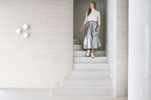 Un intérieur minimaliste et contemporain mettant en avant la collection Pure d’Omexco, référence PU251, nommée Delicate. Ce papier peint raffiné présente un raffia fin sublimé par des fils métalliques subtils, combinant une texture naturelle avec une touche d’éclat pour une esthétique sophistiquée. Une femme descend un escalier blanc vêtue d’un pull à col roulé crème et d’une jupe métallisée argentée, apportant une élégance moderne à cet espace serein. L’arrière-plan accentue l’ambiance paisible avec sa texture discrète mais complexe, idéal pour les intérieurs minimalistes valorisant le luxe subtil et l’artisanat.