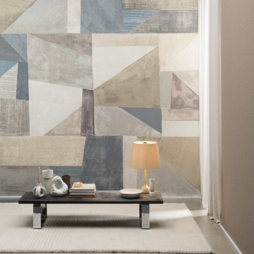 Moonstone d'Omexco est une collection somptueuse de revêtements muraux texturés, doucement irisés comme la pierre de lune. Sur cette photo, nous voyons un mur d'accent avec un art abstrait XXL imprimé sur du mica fin. Le motif géométrique aux lignes diagonales est coloré, sans être envahissant, avec ses douces nuances de beige, blanc, gris, rose tendre et bleu non saturé. Devant le mur, nous voyons une table basse en bois avec de petits vases et d'autres décorations en céramique, ainsi qu'une pile de livres et une lampe de table.