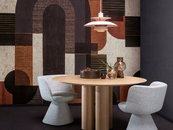 Exemple d'une salle à manger avec un revêtement mural au graphisme audacieux présentant un motif de formes géométriques dans des tons gris, noirs, orange brûlé et blancs. Ce motif panoramique est imprimé numériquement sur du liège et fait partie de la collection de revêtements muraux "Casca" d'Omexco. Au centre de la pièce se trouve une table à manger ronde en bois au-dessus de laquelle est suspendue une lampe blanche. Des éléments décoratifs tels que de petits vases en verre et un bol rempli de noix sont placés sur le plateau de la table. Deux chaises de salle à manger d'un gris doux sont disposées autour de la table. L'esthétique générale est moderne avec une touche de chaleur et de texture.