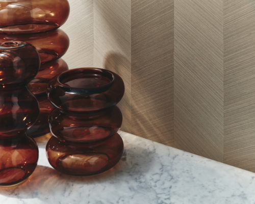 Gros plan sur une luxueuse surface en marbre sur laquelle sont disposés trois vases bruns en verre. À l'arrière-plan, la collection de revêtements muraux en marqueterie Imperialis d'Omexco constitue une toile de fond étonnante, mettant en valeur la beauté subtile d'un placage de bois fin dans un motif à chevrons, incrusté à la main de manière experte. La lumière du jour éclaire le mur d'une douce lumière, rehaussant l'élégance de la scène.