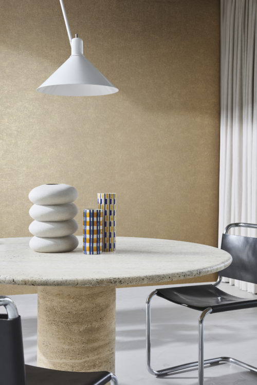 Une salle à manger contemporaine avec un revêtement mural intissé imprimé de la collection "Edition" d'Omexco, présentant un motif géométrique très subtil. Le mur a une apparence texturée beige chaude. Le décor comprend une table ronde en pierre avec une base cylindrique, décorée d'un vase sculptural blanc et de deux grands vases à motifs en bleu, jaune et blanc. Autour de la table, des chaises noires avec des cadres en chrome. Une lampe suspendue blanche est accrochée au-dessus de la table et des rideaux de couleur claire sont visibles à droite.