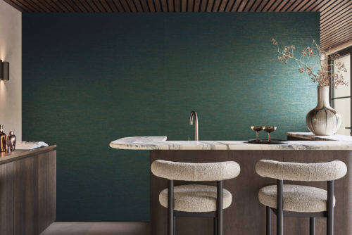 Une cuisine avec des armoires en bois sombre à gauche et un îlot de cuisine, également en bois sombre, avec un plan de travail en marbre et un robinet en cuivre. Deux chaises de bar confortables avec un tissu bouclé blanc cassé sont positionnées à l'îlot. À droite, un grand vase en céramique avec une branche décorative se trouve sur le plan de travail en marbre. Une porte en verre avec un cadre en acier noir est visible sur le côté droit de la photo. En arrière-plan, les revêtements muraux en textile de la collection Café Society d'Omexco dans des nuances de bleu sarcelle avec des touches d'or. Ce mélange harmonieux de textures et de couleurs douces crée une profondeur visuelle et un rythme vibrant.