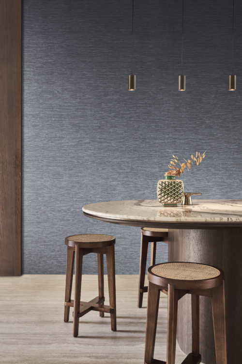 Une cuisine avec un sol en parquet naturel et un îlot de cuisine brun foncé avec un plan de travail en marbre entouré de tabourets assortis. Au-dessus de l'îlot pendent trois suspensions noires et élégantes. Sur le mur derrière l'îlot de cuisine, on voit la collection de revêtements muraux en textile Café Society dans des nuances de bleu denim. Cette collection est une réinterprétation de l'un des classiques les plus emblématiques d'Omexco, incarnant l'essence luxueuse de la soie à travers des fibres textiles méticuleusement imprimées.