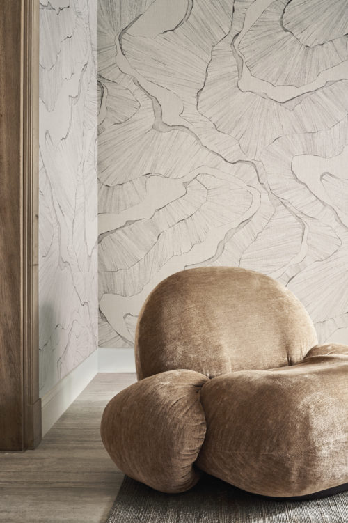 Une photo de décoration de salon avec un fauteuil rond et doux de couleur marron à l'avant, placé sur un tapis gris. Sur le mur, un design panoramique de la collection Café Society d'Omexco dans des nuances de blanc cassé. Le design fait une forte impression avec sa représentation de structures organiques, imprimées numériquement sur du lin fin.